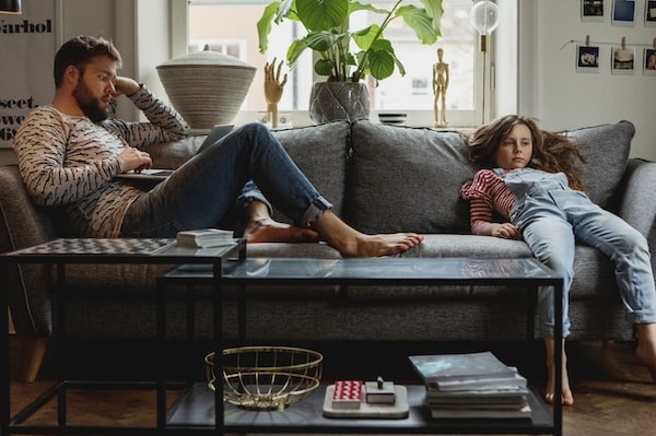 A man and a young girl lounging on a couch in a living room, both appearing relaxed and pensive, possibly discussing the cure for boredom.