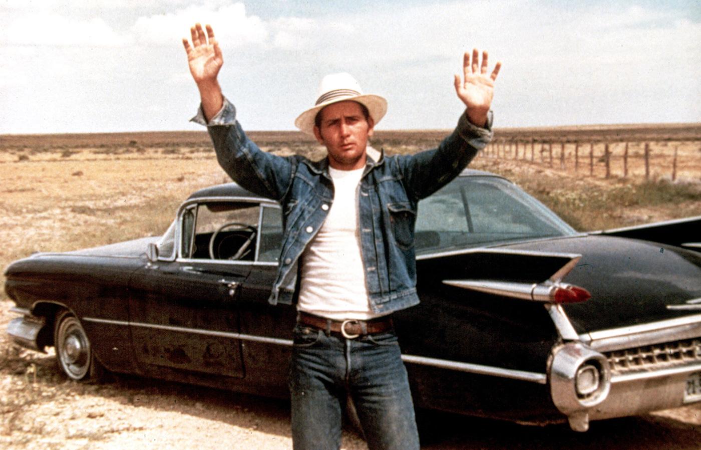 A man in a hat is standing next to a classic car, showcasing his style.