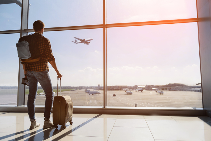 A man with a backpack looking out of an airport window, possibly booking a flight.