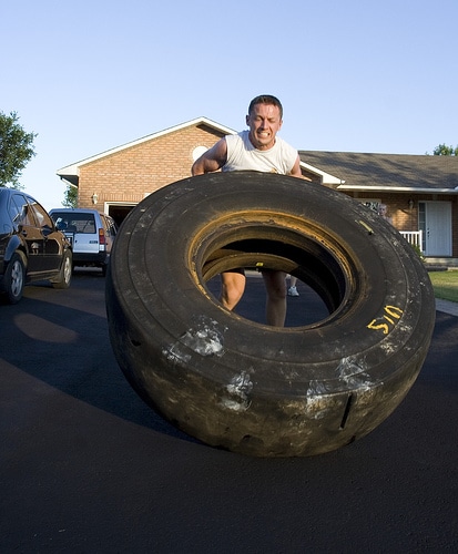 A man using DIY gym equipment to save money by lifting a large tire in front of a house.