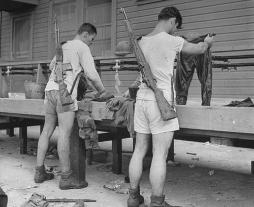 Two men in shorts standing on a bench with rifles.