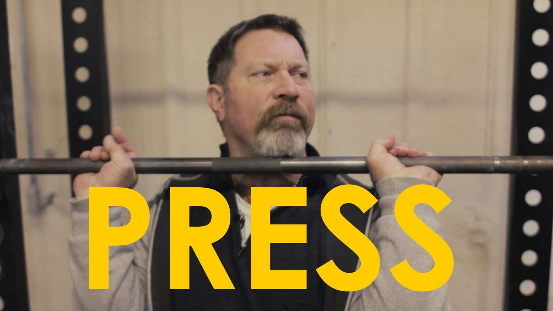 A man holding a barbell with the words press on it is demonstrating an overhead press in the video.