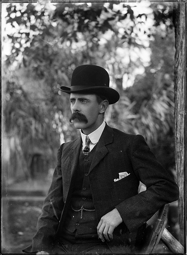 A man in a suit and hat, with a mustache, sitting on a bench.