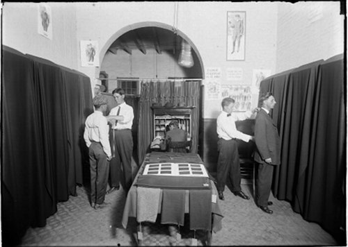 A group of men standing in front of a room with a curtain, discussing clothing alterations.