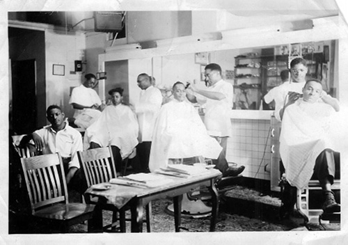 A black and white photo of men in a barber shop, getting their hair cut by a pick.