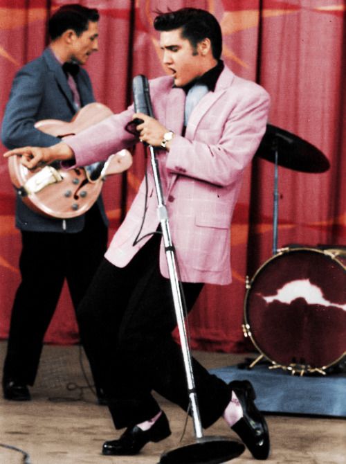 A man wearing a pink suit is singing into a microphone, resembling Elvis Presley.