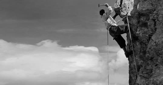 A person in climbing gear is embracing the challenge of ascending a steep rock face, using a hammer to drive in a piton for securing their rope.