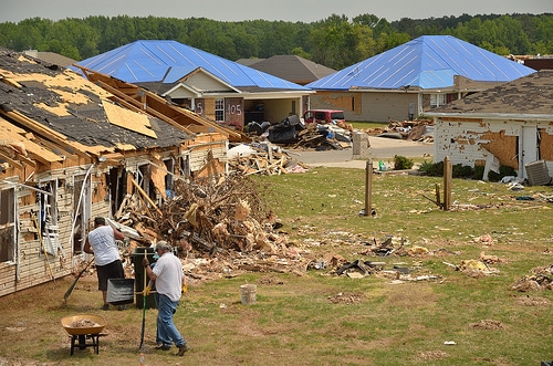 Two volunteer men standing in front of a house that has been destroyed by a tornado, providing hands-on disaster relief assistance.